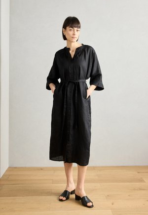 Black linen dress with a button-down front, rolled sleeves, and a tied waist. Paired with black open-toe mules. Wood floor background.