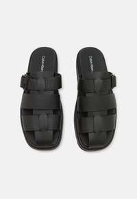 Juodi odiniai šlepetės su austa dirželio konstrukcija, sagties akcentu ir tekstūruotu paviršiumi. "Calvin Klein" logotipas matomas ant įdėklo.