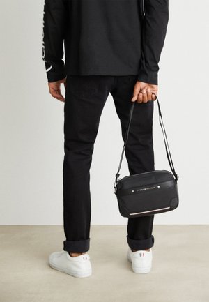 Homme portant un pantalon noir et une chemise à manches longues, tenant un sac bandoulière noir Tommy Hilfiger derrière son dos, avec des baskets blanches.