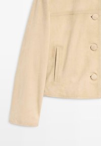 Beige ruskindsjakke med ærmer og front med stofovertrukne knapper samt en lodret slidslomme på hvid baggrund.