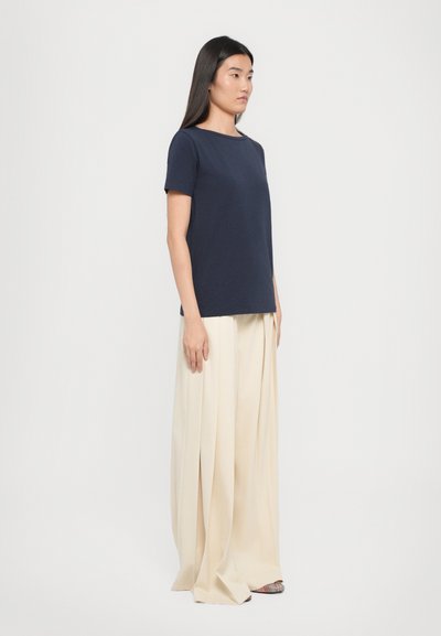 Donna in piedi di lato che indossa una maglietta corta blu navy con pantaloni a gamba larga plissettati color crema e scarpe a punta a righe.