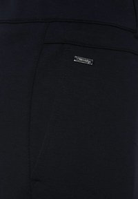 Black stretch fabric trousers featuring a front pocket and a small metallic logo accent on the side. Smooth texture with minimal stitching details.