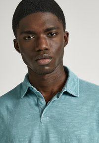 Ljusblå pikétröja i mjukt tyg med en strukturerad krage och två knappar vid halsen, visad på en modell med len hy.