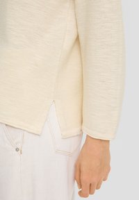 Beiger Strickpullover mit gerippter Textur, rundem Halsausschnitt und seitlichen Schlitzen. Kombiniert mit heller Denimhose mit sichtbaren Nähten.
