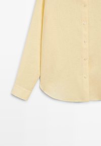 Chemise jaune clair à manches longues avec boutons aux poignets, montrée de la taille jusqu’à la manche gauche sur fond blanc.
