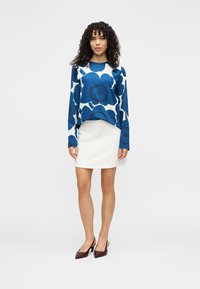 Pulover albastru cu model floral și mâneci lungi, asortat cu o fustă mini albă și pantofi cu toc ascuțit de culoare roșu închis. Texturi netede peste tot.