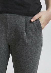 Pantalon gris à motifs avec plis à l'avant, poches latérales et texture lisse, associé à une chemise en coton noire sur le bord supérieur.