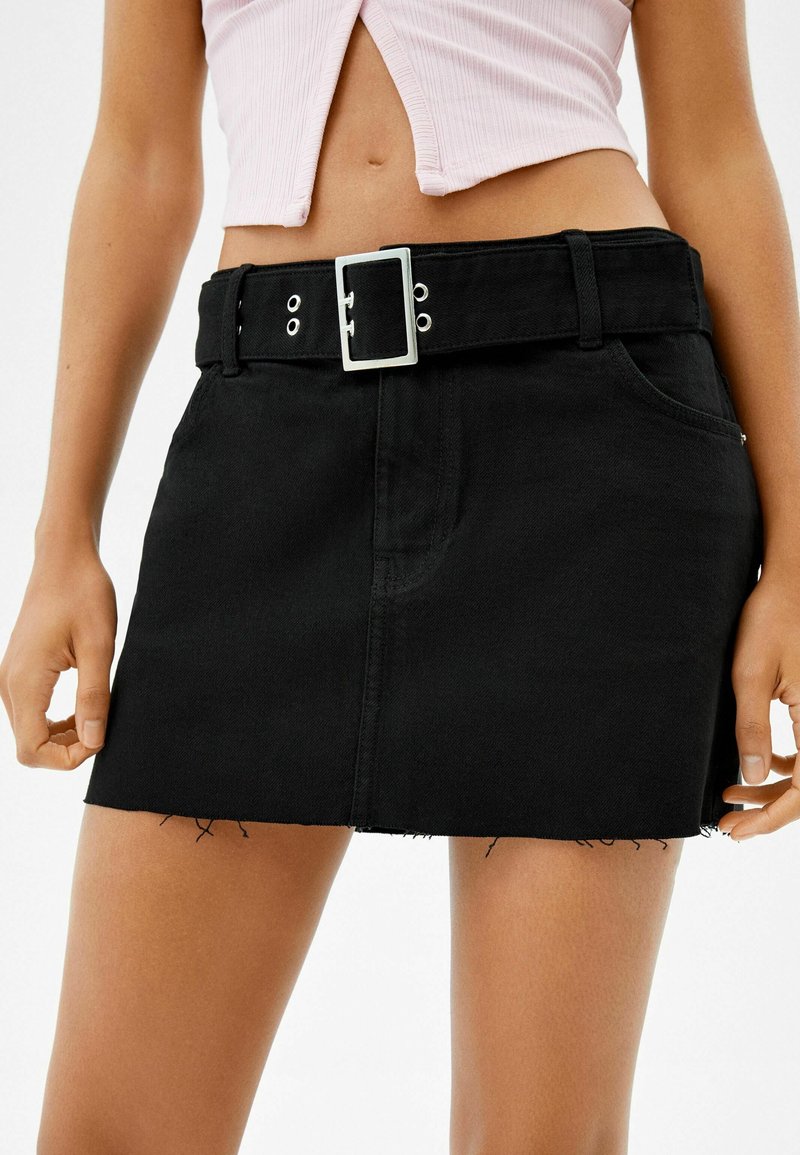 Svart denimminikjol med fransad kant, utrustad med ett brett bälte med silverspänne och två framfickor.