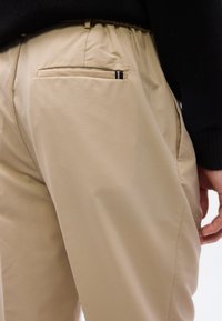 Μπεζ παντελόνι με πίσω τσέπη welt που έχει μια μικρή κόκκινη, λευκή και μαύρη ριγέ ετικέτα, συνδυασμένο με μανίκι μαύρου πουλόβερ που φαίνεται μερικώς.