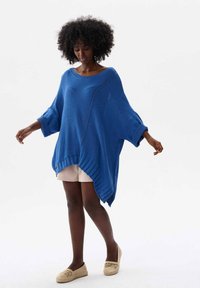 Losse blauwe gebreide trui met een brede halslijn, lange mouwen en een asymmetrische zoom. Gecombineerd met beige shorts en lichte instappers.