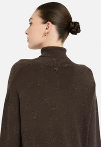 Bruine gebreide trui met een hoge kraag, voorzien van verspreide zilveren spekjes, een drop shoulder ontwerp en een klein logo op het gebied van de achterkant van de nek.