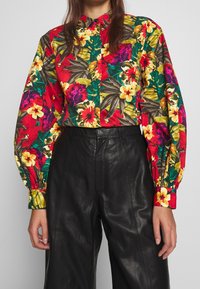 Chemise à boutons à motifs floraux avec des teintes vives de rouge, vert et jaune, dotée de manches longues bouffantes. Associée à un pantalon en cuir noir taille haute.