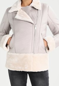 Chaqueta con cremallera de color gris claro, con cuello y puños de piel sintética. Presenta un forro texturizado y un dobladillo de color crema suave. Diseño ajustado.