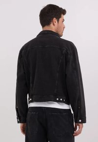 Sort denimjakke med et cropped design, synlig syning og metaltrykknapper i kanten. Har en klassisk krave og detaljer ved bagsømmen.
