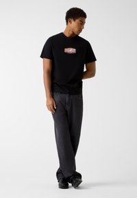 Czarny bawełniany t-shirt z naszywką z logo w kolorze czerwono-białym. Połączony z ciemnoszarymi dżinsami i czarnymi butami, tworząc monochromatyczny wygląd.