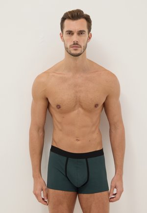 Shirtless man with short brown hair wearing dark green boxer briefs with black waistband standing against a plain light wall.