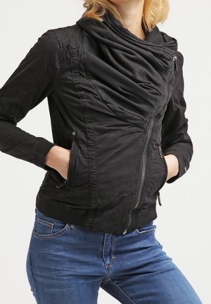 Frau trägt eine figurbetonte schwarze Jacke mit einem drapierten Kragen und diagonalem Reißverschluss, Hände in den Taschen, kombiniert mit blauen Jeans.