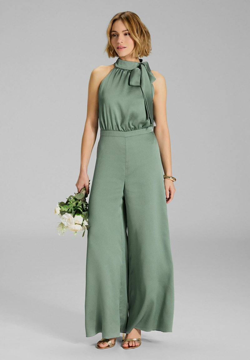 Groene wijde jumpsuit met een hoge, strikhals. Gemaakt van textuurstof, met een aansluitende bovenkant en vloeiende benen. Houdt een boeket vast.