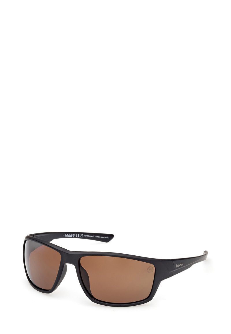 Lunettes de soleil en plastique noir avec des verres teintés bruns, monture rectangulaire épaisse et marque Timberland sur la branche. Design moderne et épuré.