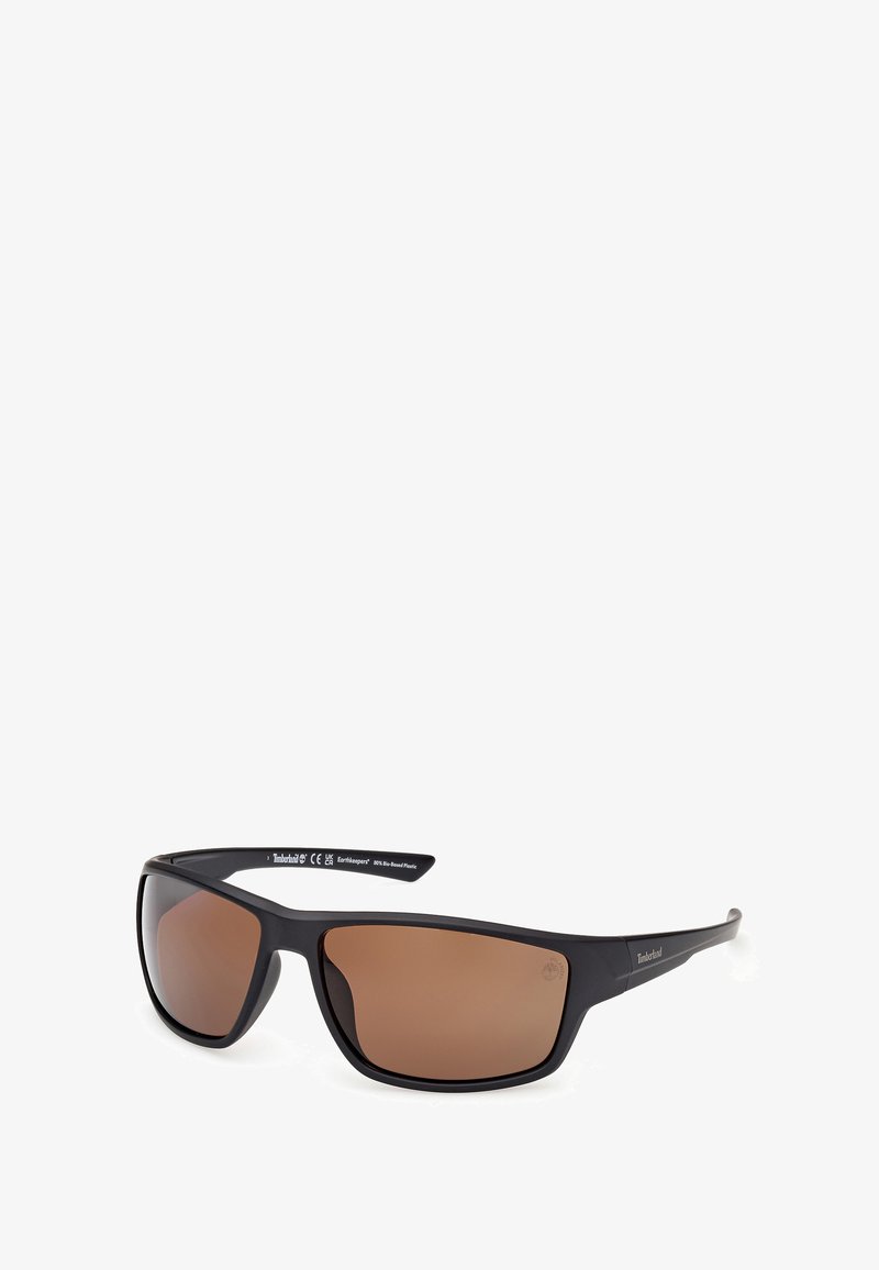 Lunettes de soleil en plastique noir avec des verres teintés bruns, monture rectangulaire épaisse et marque Timberland sur la branche. Design moderne et épuré.