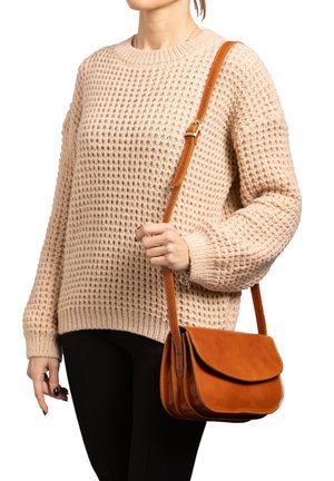Borsa a tracolla in pelle marrone con chiusura a patta, bordi cuciti e una sola tracolla regolabile, tenuta in mano contro un maglione a trama.
