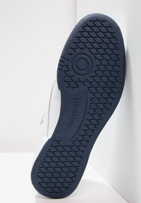 Semelle d'une basket blanche avec un motif en caoutchouc bleu marine comportant des cercles et des losanges, avec le logo Reebok embossé au centre.