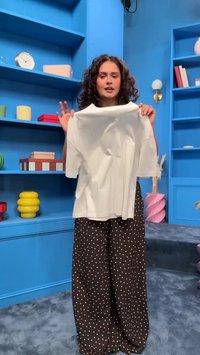 Frau mit lockigem Haar hält ein schlichtes weißes Kurzarmshirt und steht in einem blauen Raum mit Regalen, auf denen dekorative Gegenstände stehen.