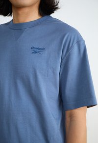 Tricou din bumbac albastru, cu mâneci scurte, având un guler rotund și un logo brodat Reebok de culoare albastru mai închis pe partea stângă a pieptului.