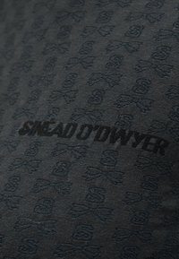 Μαύρο ύφασμα με επαναλαμβανόμενο ανάγλυφο σχέδιο κρανίου και σταυρωμένων οστών και το έντονο κείμενο «SNEAD O'DWYER» σε μαύρο στο κέντρο.