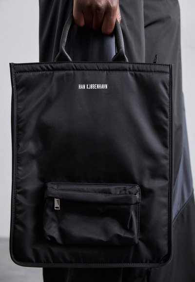 Black nylon tote bag with two short handles, front zip pocket, and branding "HAN KJØBENHAVN" in white text. Smooth texture, rectangular shape.