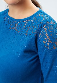 Maglione blu con un collo in pizzo a motivi floreali, arricchito da piccole strass lungo il corpo per una texture e un dettaglio raffinati.