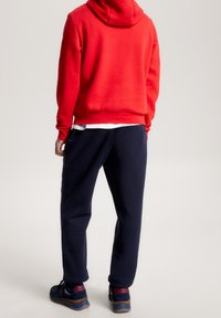 Felpa rossa con cappuccio e vestibilità comoda, abbinata a pantaloni della tuta blu navy. Sneakers nere con dettagli blu e rossi. Design semplice e confortevole.