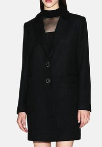 Mujer con un abrigo negro de longitud media y doble botonadura sobre una blusa negra transparente con cuello, sobre un fondo claro liso.
