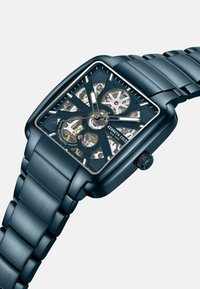 Automatische Uhr mit einem rechteckigen blauen Gehäuse, transparentem Zifferblatt, das Zahnräder zeigt, und einem passenden blauen Metallarmband mit gebürsteter Oberfläche.
