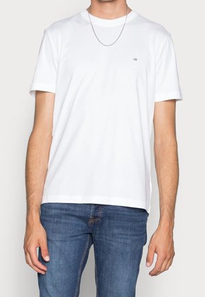 Man draagt een effen wit T-shirt met korte mouwen en een klein "ck" logo op de borst, blauwe spijkerbroek en een dun zilveren ketting.
