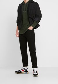 Herroutfit med en svart denimjacka med foder av fåll, mörkgrön huvtröja, svarta byxor och svarta sneakers från New Balance.