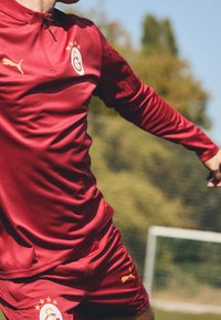 Rød trøje og shorts med en tekstureret overflade, der har høj krave, Puma-logo og klubemblem med stjerner på brystet.