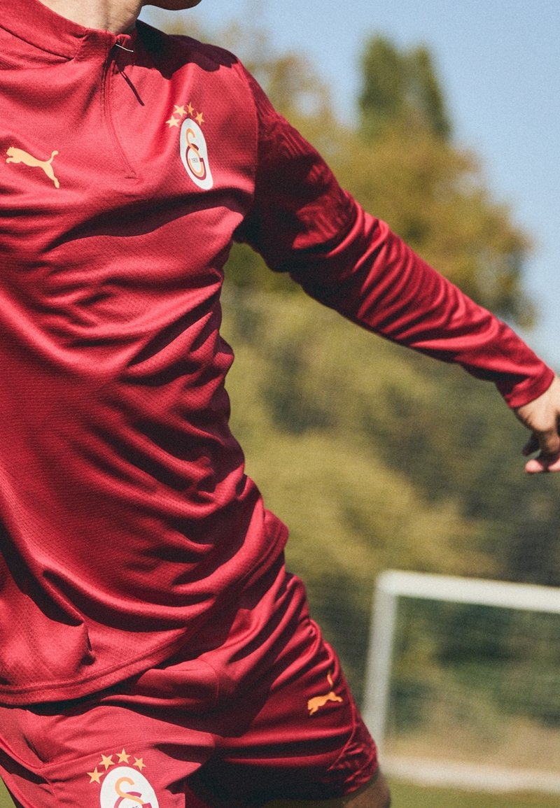 Rød trøje og shorts med en tekstureret overflade, der har høj krave, Puma-logo og klubemblem med stjerner på brystet.