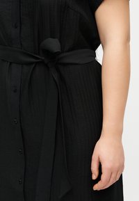 Μαύρο υφασμάτινο φόρεμα με κουμπιά και δέσιμο στη μέση, εμφανίζεται από τη μέση έως το μέσο του μηρού, με το δεξί χέρι χαλαρό δίπλα στο σώμα.