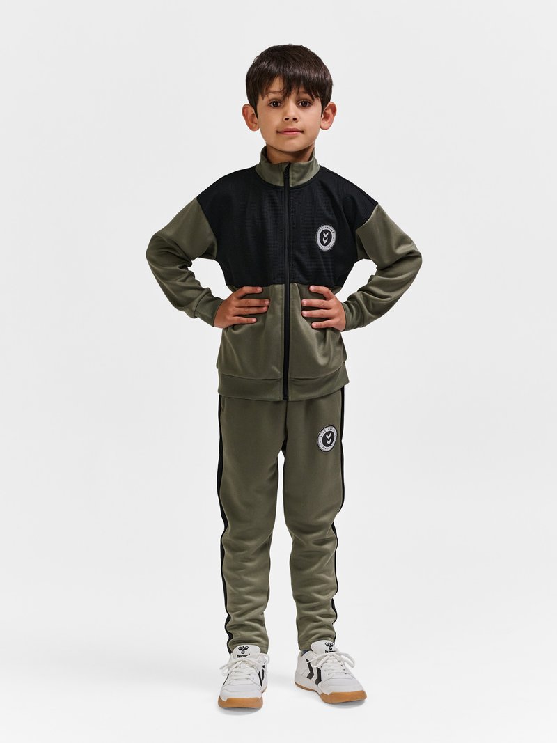 En ung dreng står med hænderne på hofterne iført en olivengrøn og sort træningsdragt med hvide sneakers mod en ensfarvet lys baggrund.