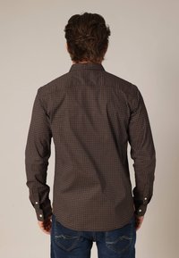 Langarmshirt van donkerbruin stof, met een klein geometrisch patroon in rood en crème. Met knoopsluiting bij de mouwen en een gespreide kraag.