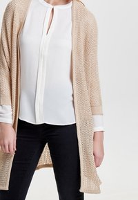 Světle hnědý pletený cardigan s otevřeným předním dílem a tříčtvrtečními rukávy, nošený přes bílou blůzu, doplněný černými přiléhavými džíny.