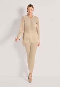 Beige Langarmbluse mit Rundhalsausschnitt und dezenter Knopfleiste an der Vorderseite, kombiniert mit passenden schmalen Hosen und spitzen beigen Absätzen.