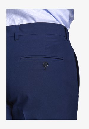 Marineblaue Anzughose mit einer letzten Gesäßtasche, die einen einzelnen schwarzen Knopf hat, getragen mit einem in die Hose gesteckten hellblauen Hemd.