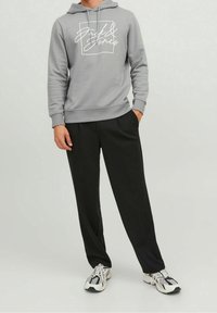 Sudadera gris con un logotipo gráfico blanco, combinada con pantalones negros con plisados y zapatillas multicolores, creando un atuendo casual.