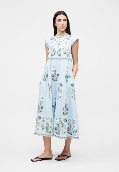 Γυναίκα που φοράει ανοιχτό μπλε αμάνικο midi φόρεμα με κεντήματα λουλουδιών και μαύρες σαγιονάρες, στέκεται με τα χέρια στις τσέπες, κοιτάζοντας προς τα πλάγια.