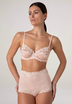 Donna che indossa un reggiseno in pizzo rosa chiaro abbinato e pantaloncini a vita alta, in piedi con le mani sui fianchi su uno sfondo semplice.