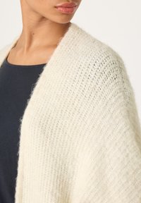 Stickad sjal i mjukt, krämfärgat tyg med ett texturerat mönster och avslappnad hängning. Närbild på halsringning och axel.