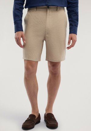 Mann trägt beige knielange Shorts, marineblaues Langarmshirt und braune Loafers, steht vor einem schlichten hellen Hintergrund.