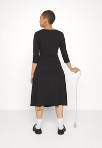 Femme aux cheveux courts portant une robe noire et des chaussures, tenant une canne blanche, debout et tournée dos à l'appareil photo.